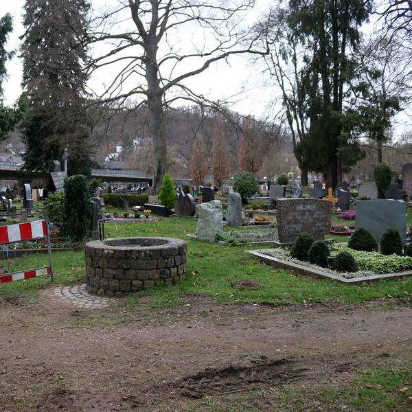 Das Foto zeigt den Friedhof, im Vordergrund eine Wasserzapfstelle, daneben ein Bauzaun, mit dem ein Weg abgesperrt wird.