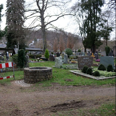 Das Foto zeigt den Friedhof, im Vordergrund eine Wasserzapfstelle, daneben ein Bauzaun, mit dem ein Weg abgesperrt wird.