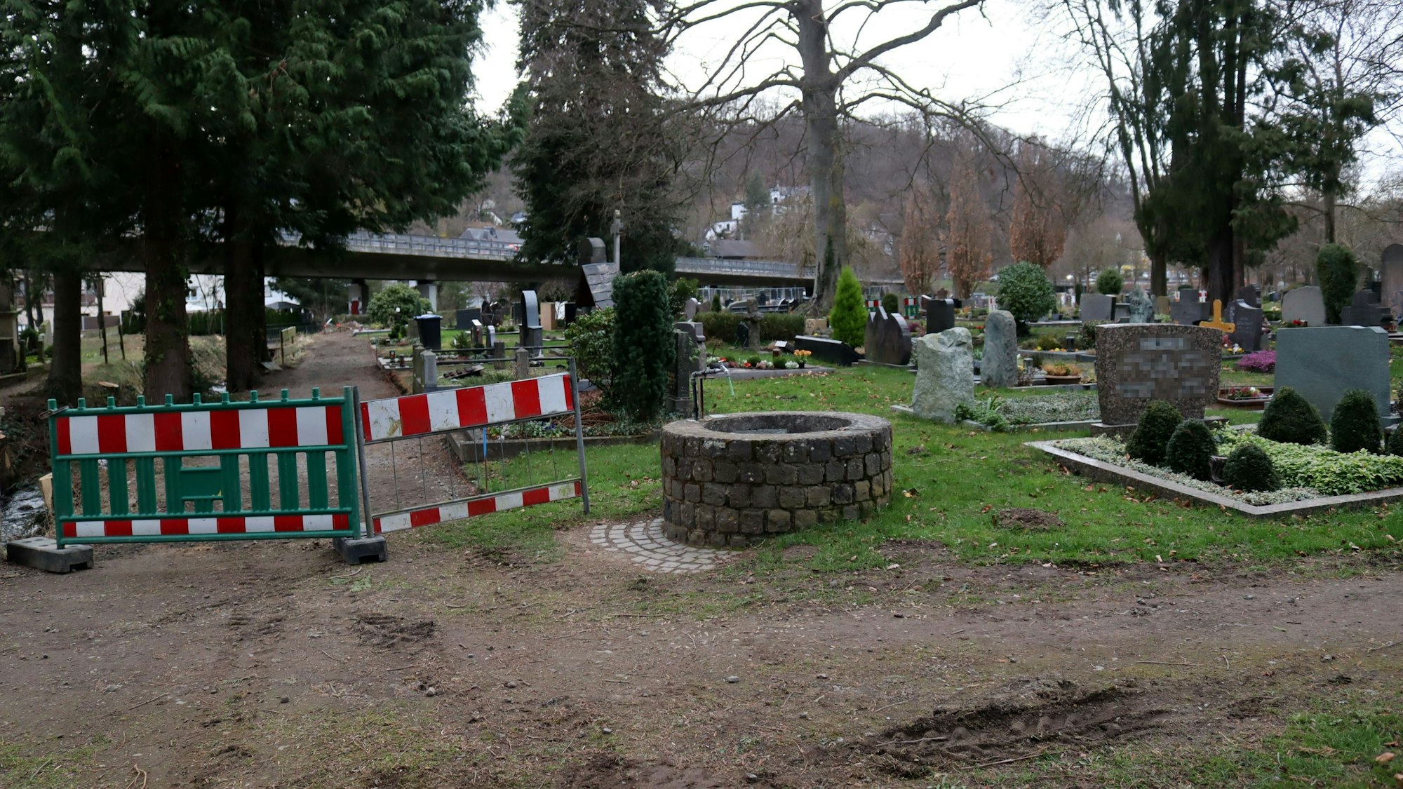 Das Foto zeigt den Friedhof, im Vordergrund eine Wasserzapfstelle, daneben ein Bauzaun, mit dem ein Weg abgesperrt wird.