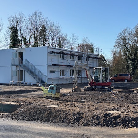 Auf dem Bild ist eine Baustelle mit Modulbauten und einem Bagger zu sehen.
