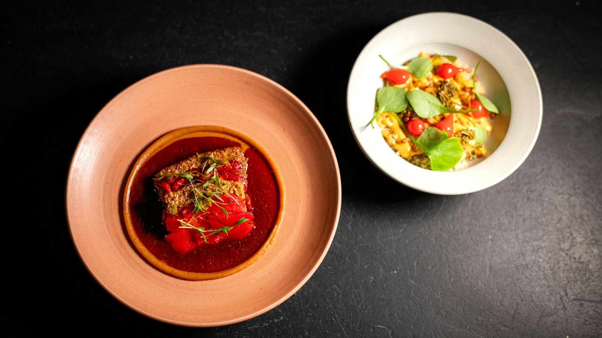 Zwei Teller sind zu sehen, einmal die Kochbanane als Karreee mit gegrillter Paprika bedeckt, sowie wie ein Schälchen mit Couscous und frischen Kräutern.