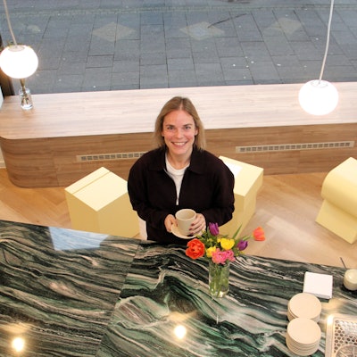 Eine Frau in schwarzer Jacke steht mit einer Kaffeetasse in der Hand vor einem Tresen.