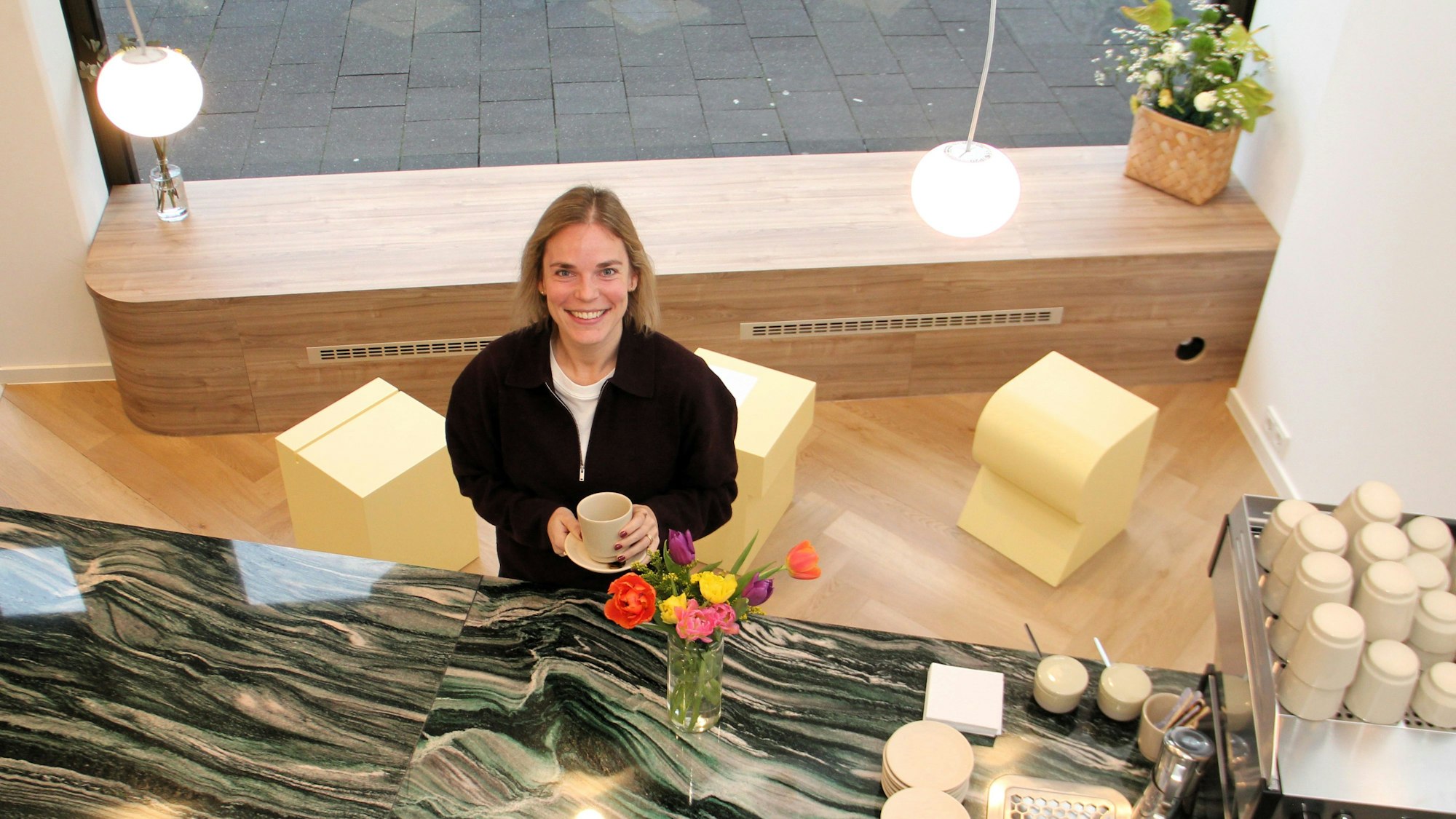 Eine Frau in schwarzer Jacke steht mit einer Kaffeetasse in der Hand vor einem Tresen.