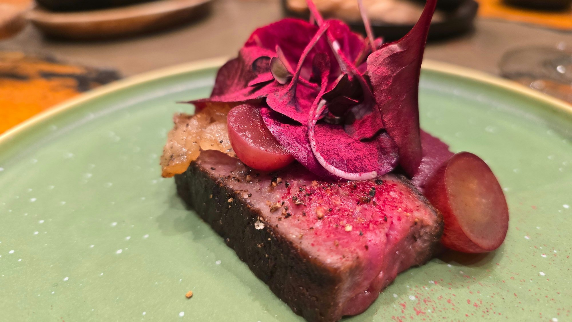 Eine Tranche rosa gebratenes Rindfleisch wird getoppt von lila Blättern, daneben liegen aufgeschnittene Trauben.