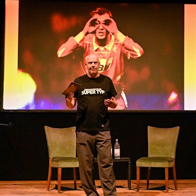 Mario Basler steht mit einer Moderatonskarte auf einer Bühne. Hinter ihm wird ein Bild aus seiner Jugend im Fußballtrikot gezeigt.