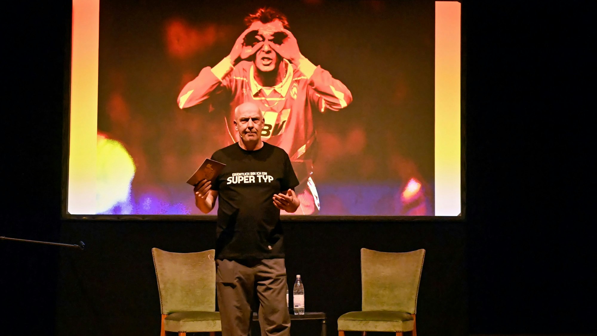 Mario Basler steht mit einer Moderatonskarte auf einer Bühne. Hinter ihm wird ein Bild aus seiner Jugend im Fußballtrikot gezeigt.