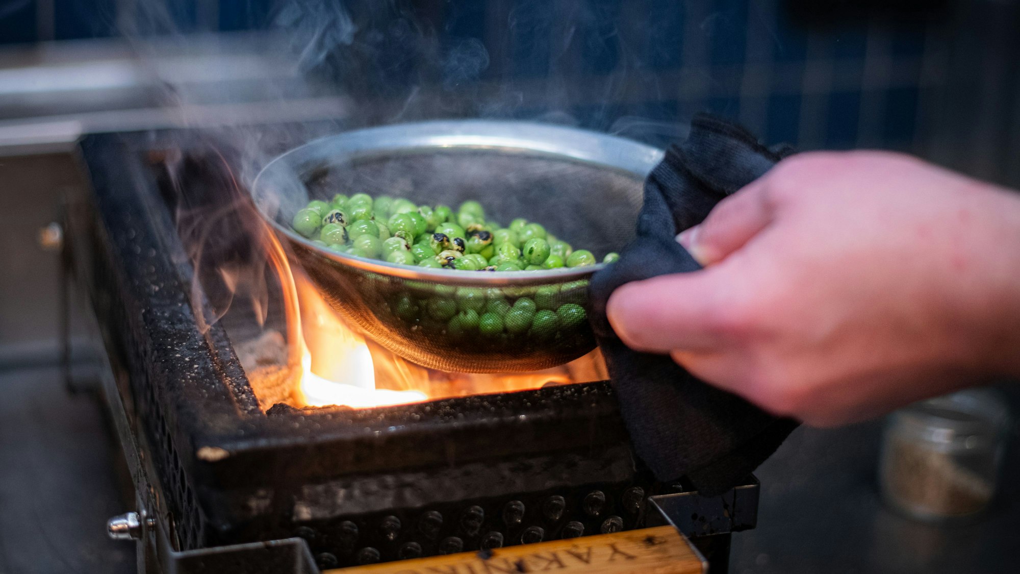 Eine Hand hält ein Sieb mit Erbsen über eine offene Flamme.