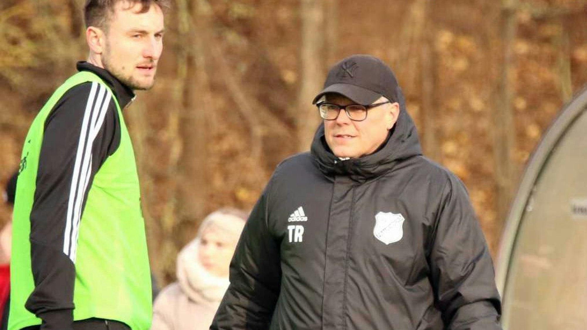 Marcel Timm trägt Kappe und dicke Jacke. Neben ihm macht sich ein Spieler warm.