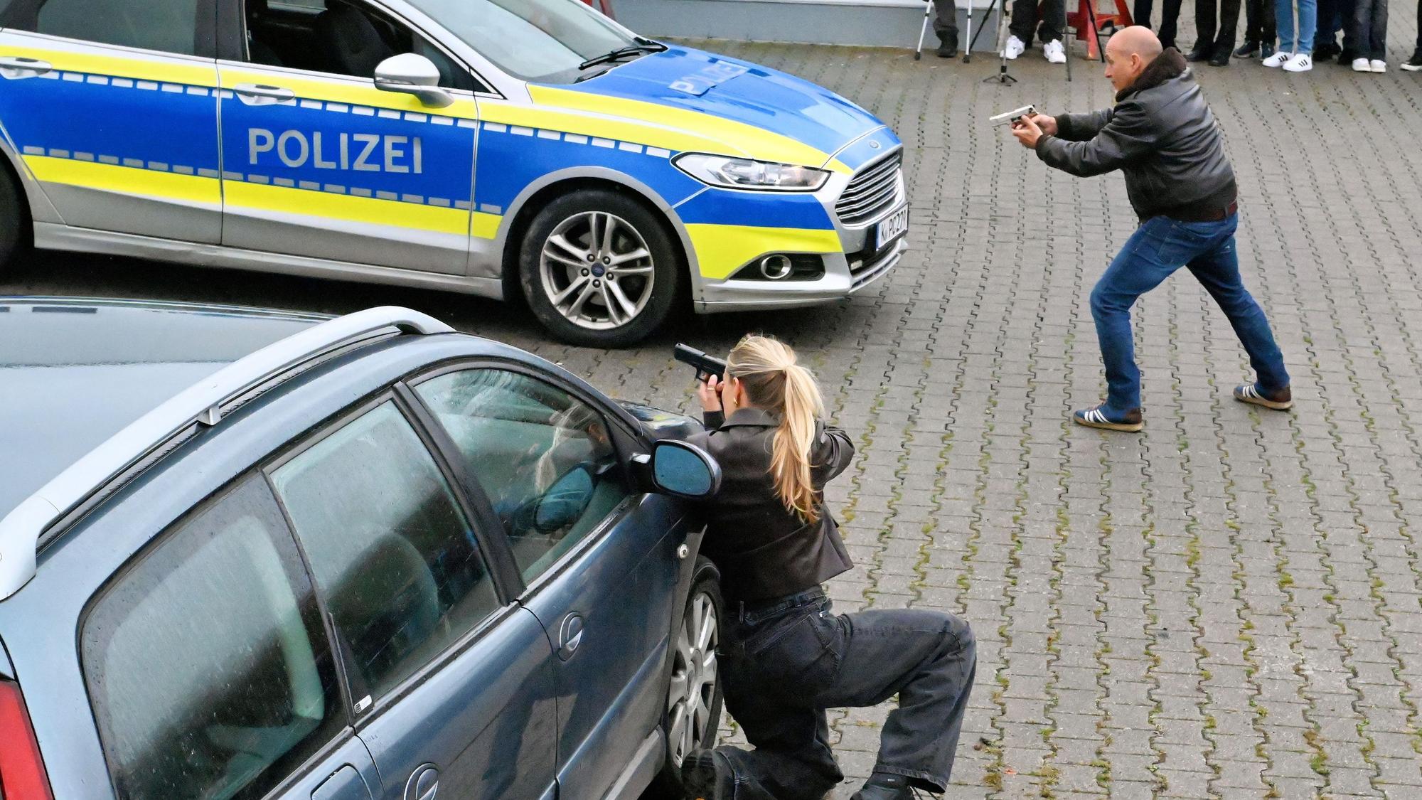 Schauspieler Erdogan Atalay und Pia Stutzenstein stehen und knien mit gezückter Waffe an einem Polizeiauto.