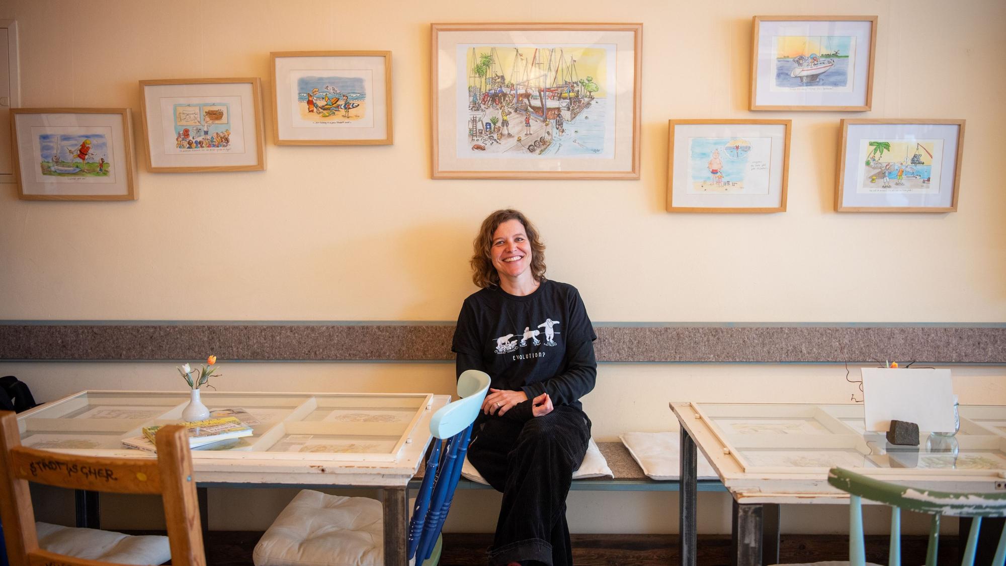 Inga Beitz-Svechtarov sitzt im Fair-Café in Zülpich vor einer Wand, an der ihre Cartoons hängen. Im Vordergrund sind Tische und Stühle zu erkennen.