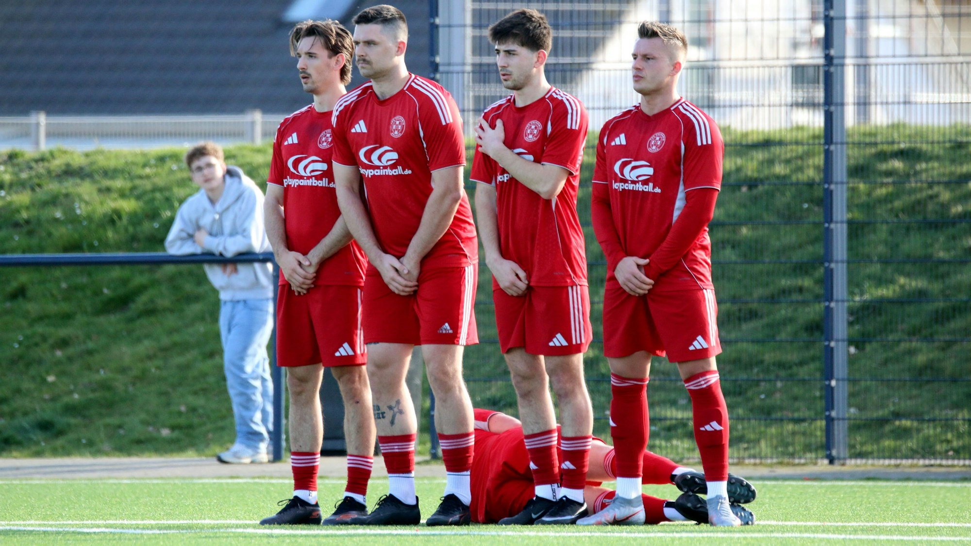 Vier Fußballer stehen in einer Mauer und halten sich die Hände vor den Schritt. Hinter der Mauer liegt ein Spieler auf dem Boden.