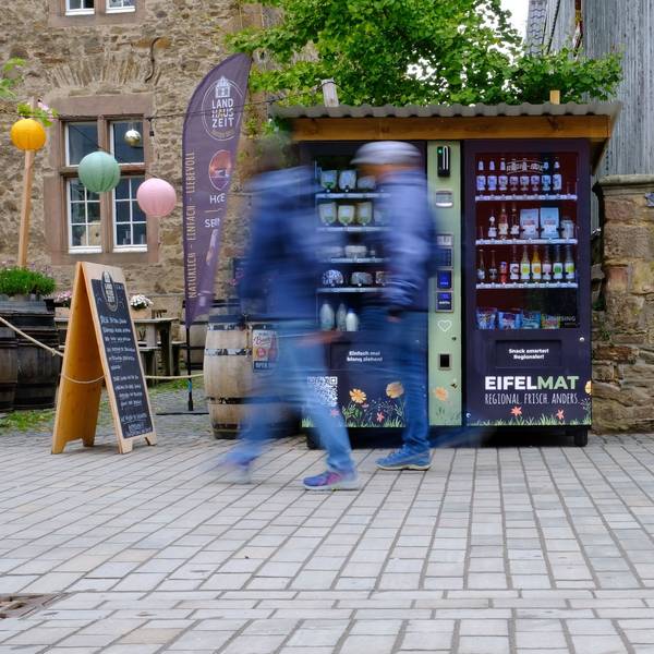 An einem Verkaufsautomaten, der in der Fußgängerzone in Bad Münstereifel steht, gehen Passanten vorbei, die bewegungsunscharf dargestellt werden.