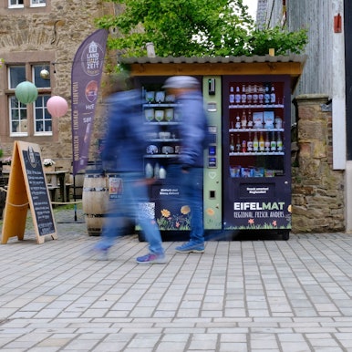An einem Verkaufsautomaten, der in der Fußgängerzone in Bad Münstereifel steht, gehen Passanten vorbei, die bewegungsunscharf dargestellt werden.