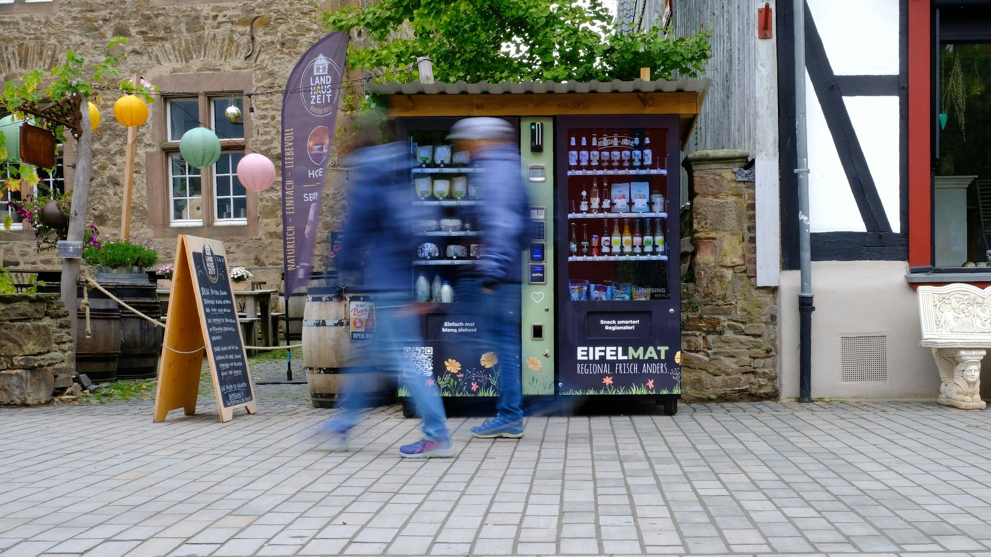 An einem Verkaufsautomaten, der in der Fußgängerzone in Bad Münstereifel steht, gehen Passanten vorbei, die bewegungsunscharf dargestellt werden.
