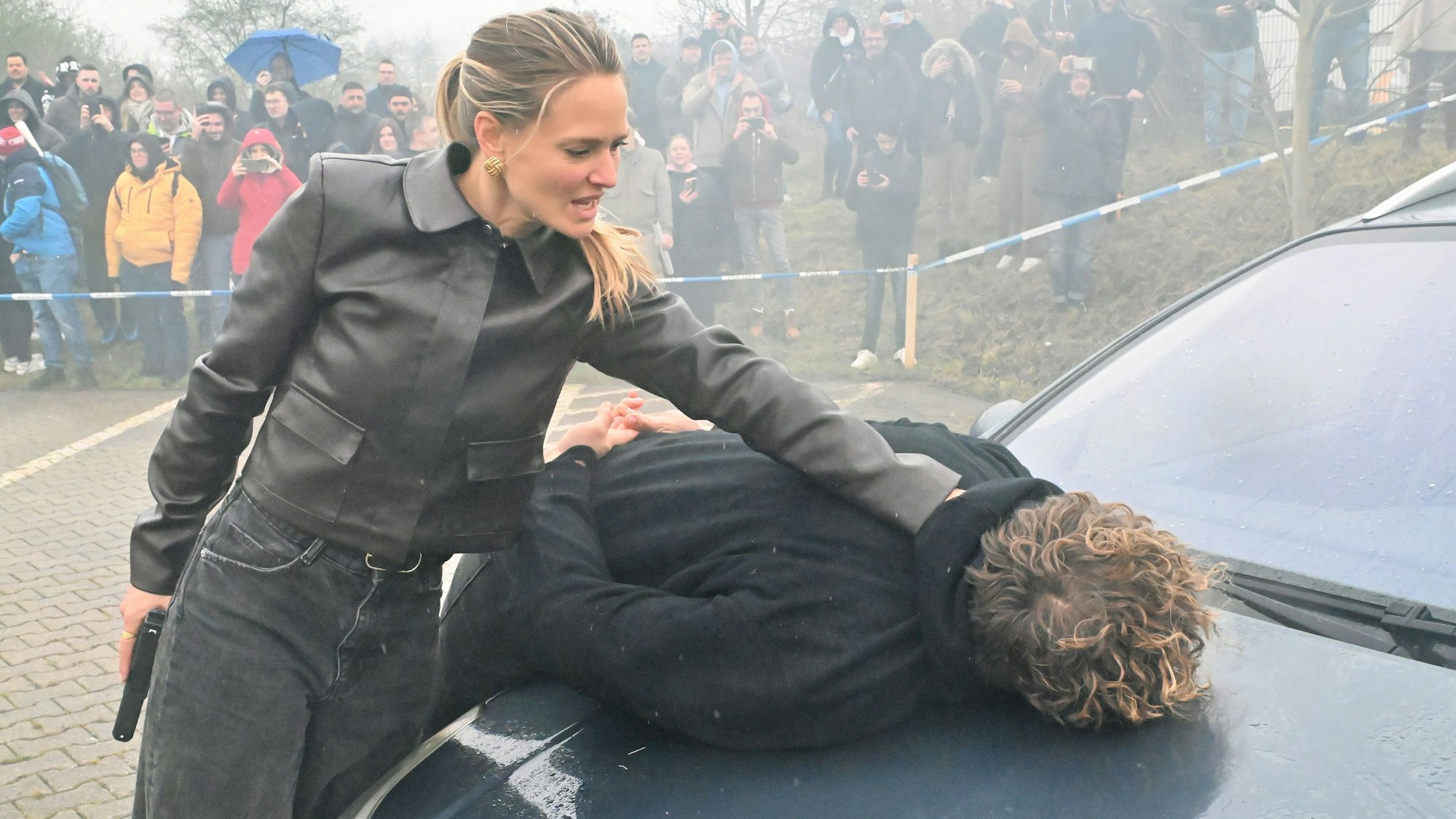 Die schwarz gekleidete Darstellerin drückt einen Mann auf die Motorhaube eines Autos.