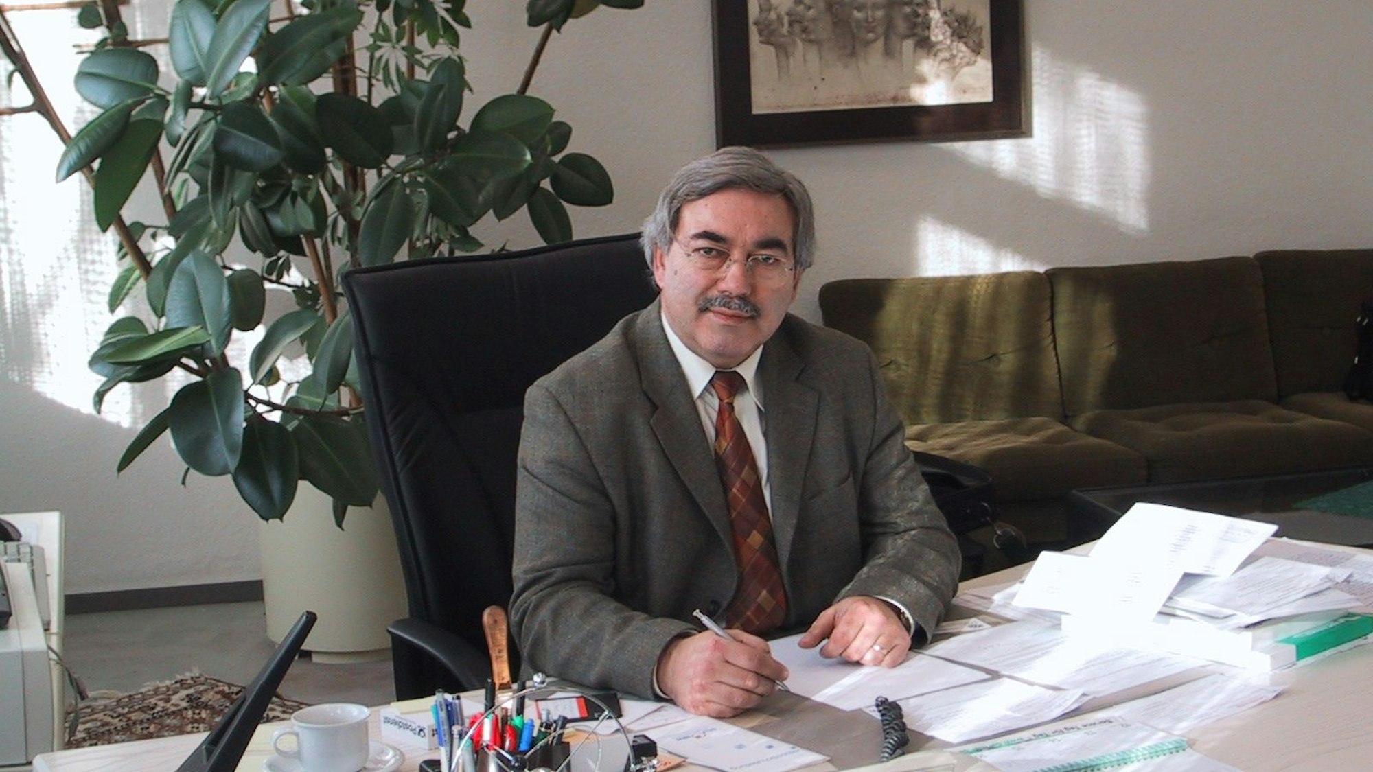 Das Bild zeigt den ehemaligen Schulleiter sitzend auf einem Bürostuhl, vor sich eine Reihe von Papieren. Er hält einen Stift in der Hand und trägt einen Anzug mit roter Krawatte.