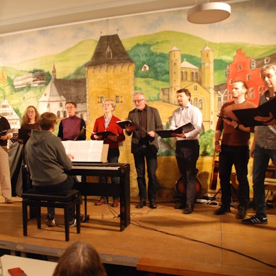 Die Sängerinnen und Sänger stehen in einer Reihe auf der Bühne. Der Dirigent sitzt vor ihnen an einem E-Piano.