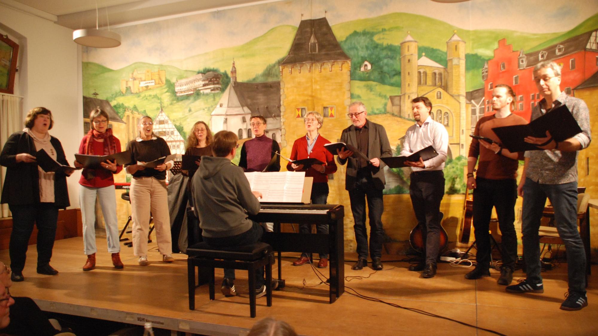 Die Sängerinnen und Sänger stehen in einer Reihe auf der Bühne. Der Dirigent sitzt vor ihnen an einem E-Piano.