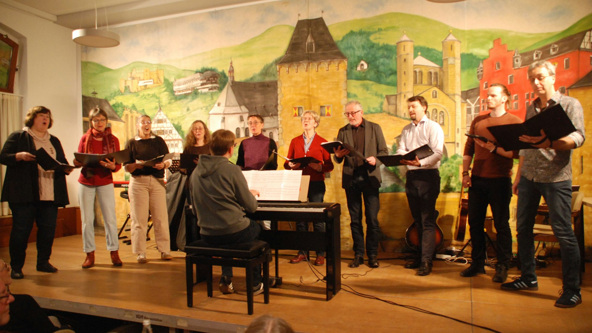 Die Sängerinnen und Sänger stehen in einer Reihe auf der Bühne. Der Dirigent sitzt vor ihnen an einem E-Piano.