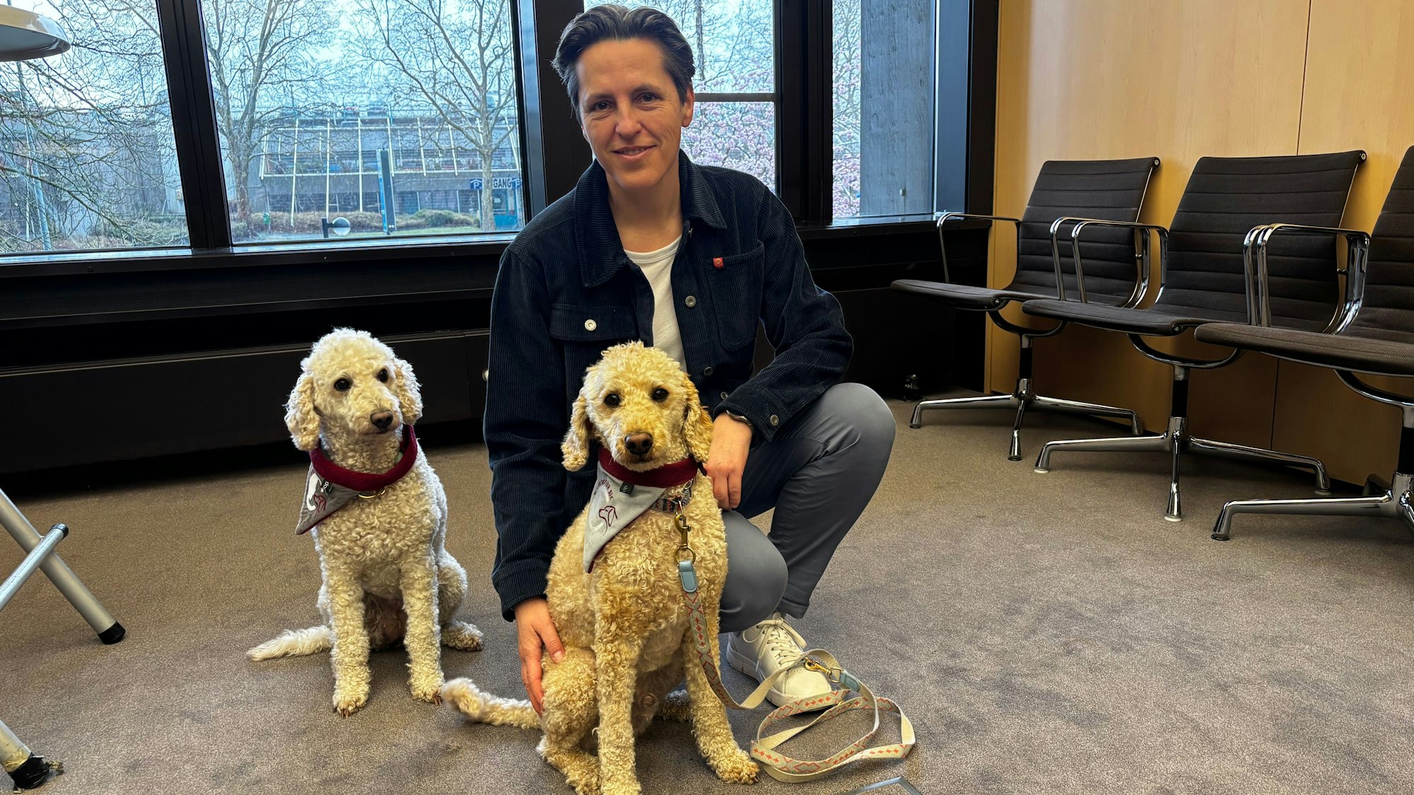 Eine Frau kniet auf der Erde und lächelt in die Kamera. Vor ihr sitzen zwei cremefarbene Pudel.