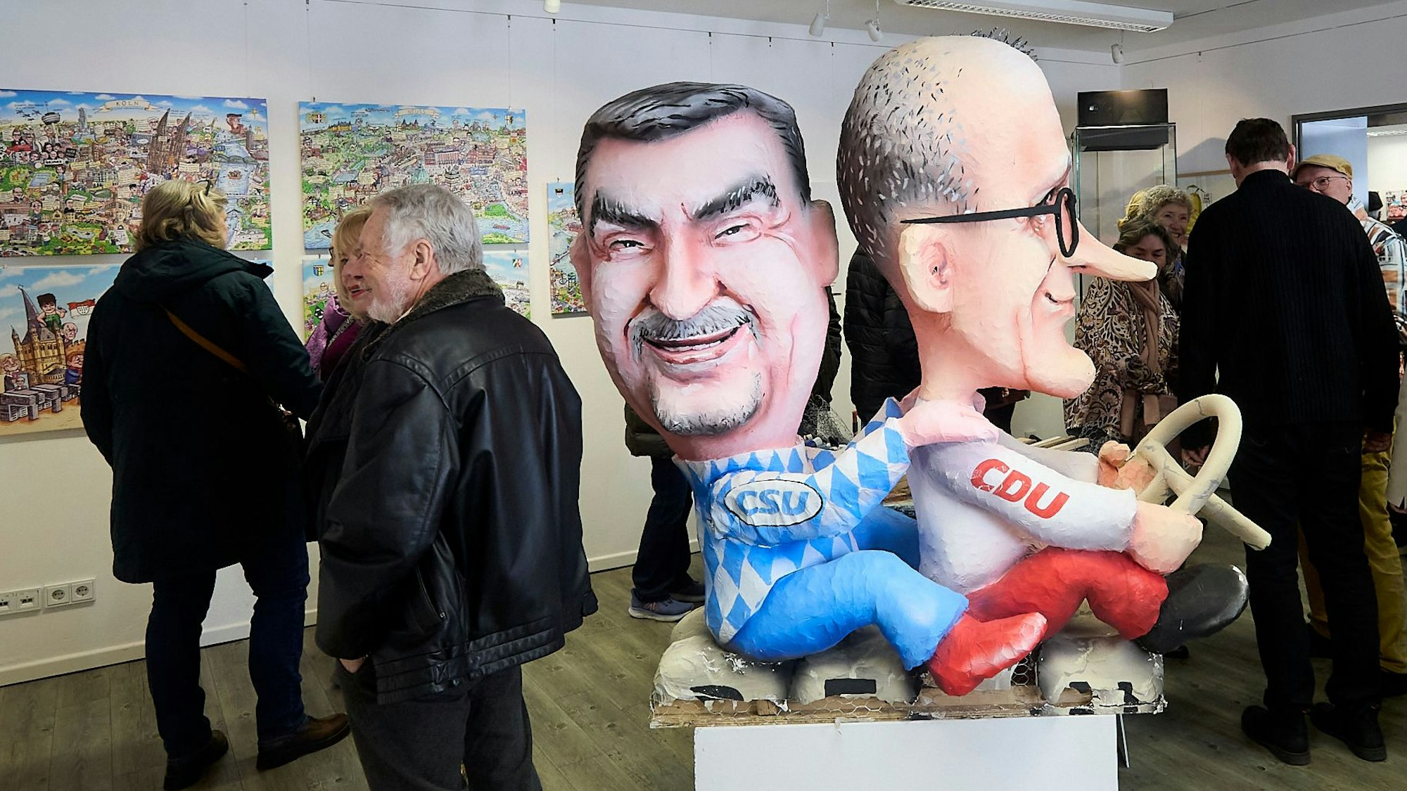 Die Figuren von Markus Söder und Friedrich Merz sitzen hintereinander, Merz hält ein Lenkrad. Im Hintergrund betrachten Menschen die Exponate.