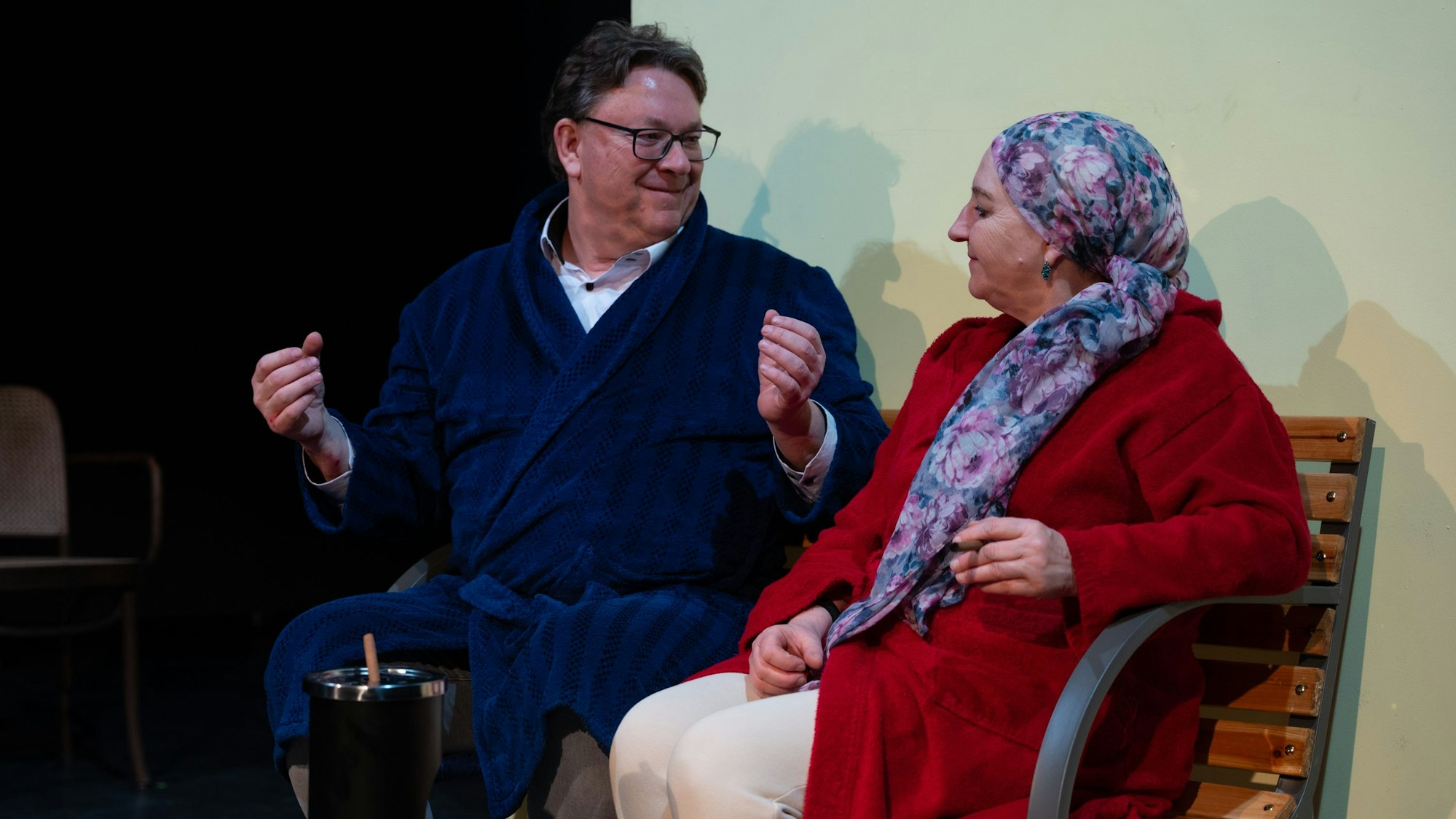 Zwei Schauspieler sitzen auf einer Bank und unterhalten sich.