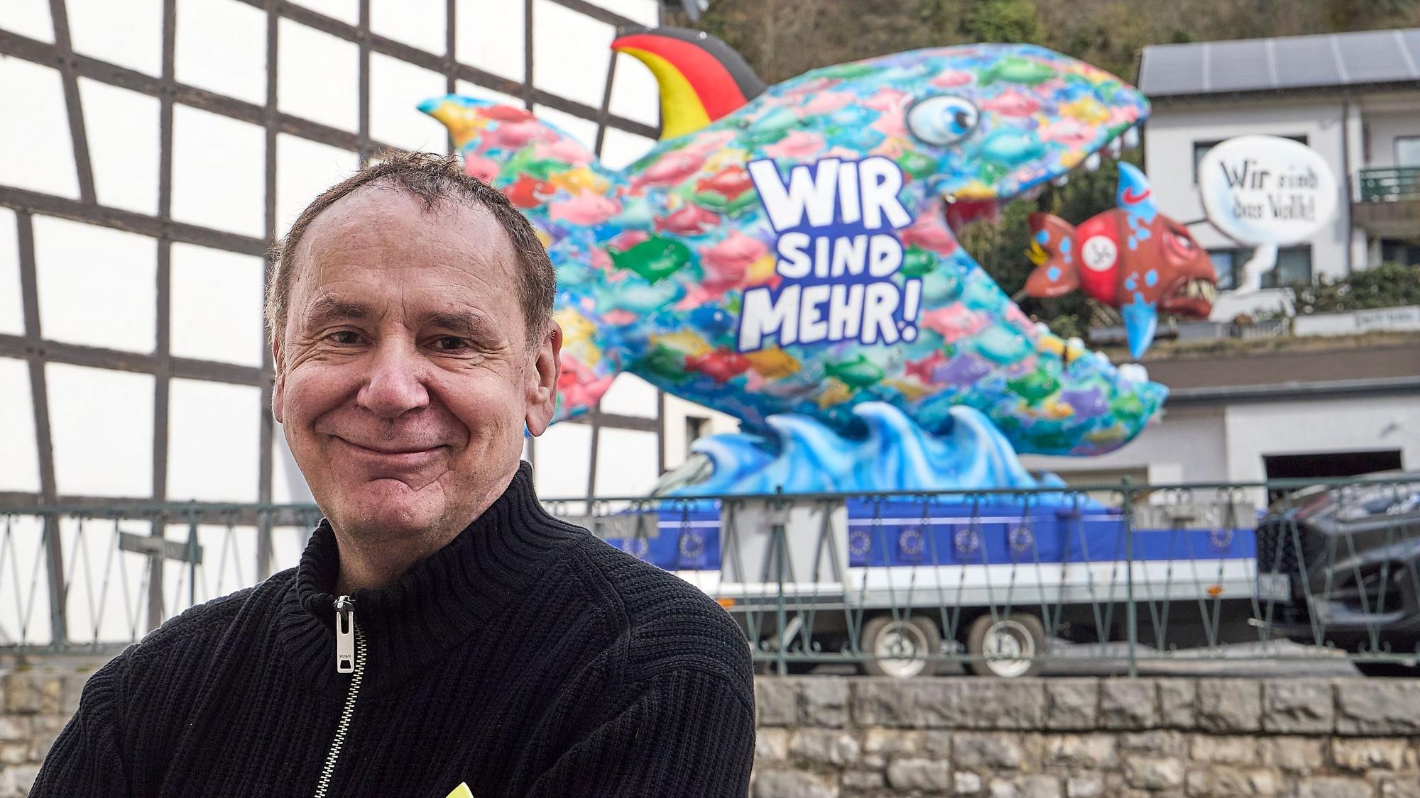Jacques Tilly stehrt vor einem seiner Karnevalswagen. Der zeigt einen großen Fisch, der einen kleinen verschluckt.