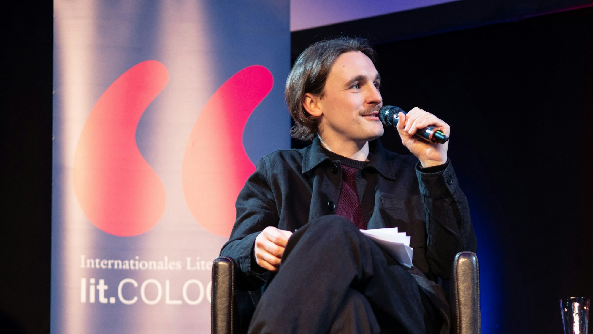 Zwei Männer sitzen, jeweils mit einem Mikrofon in der Hand, auf einer Bühne.