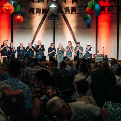 Eine Reihe Eifeler Musiker steht auf einer Bühne, die angeleuchtet wird. Vor ihnen befindet sich ein Publikum, das mit dem Rücken zur Kamera gewandt ist, im Dunklen.