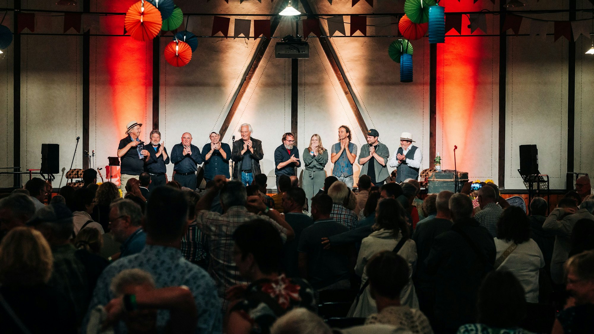 Eine Reihe Eifeler Musiker steht auf einer Bühne, die angeleuchtet wird. Vor ihnen befindet sich ein Publikum, das mit dem Rücken zur Kamera gewandt ist, im Dunklen.