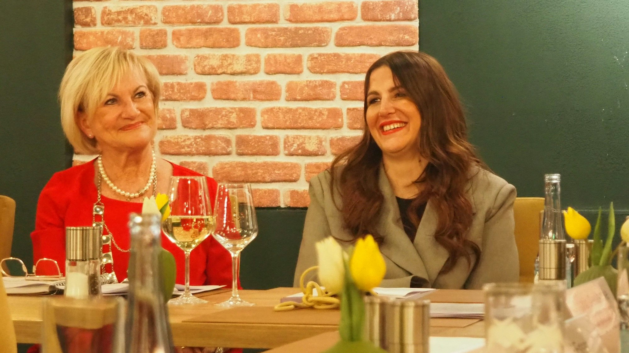 Bernadette Hein (l.) und Judith Kneuttinger sitzen an einem Tisch mit Weingläsern und Blumen darauf.