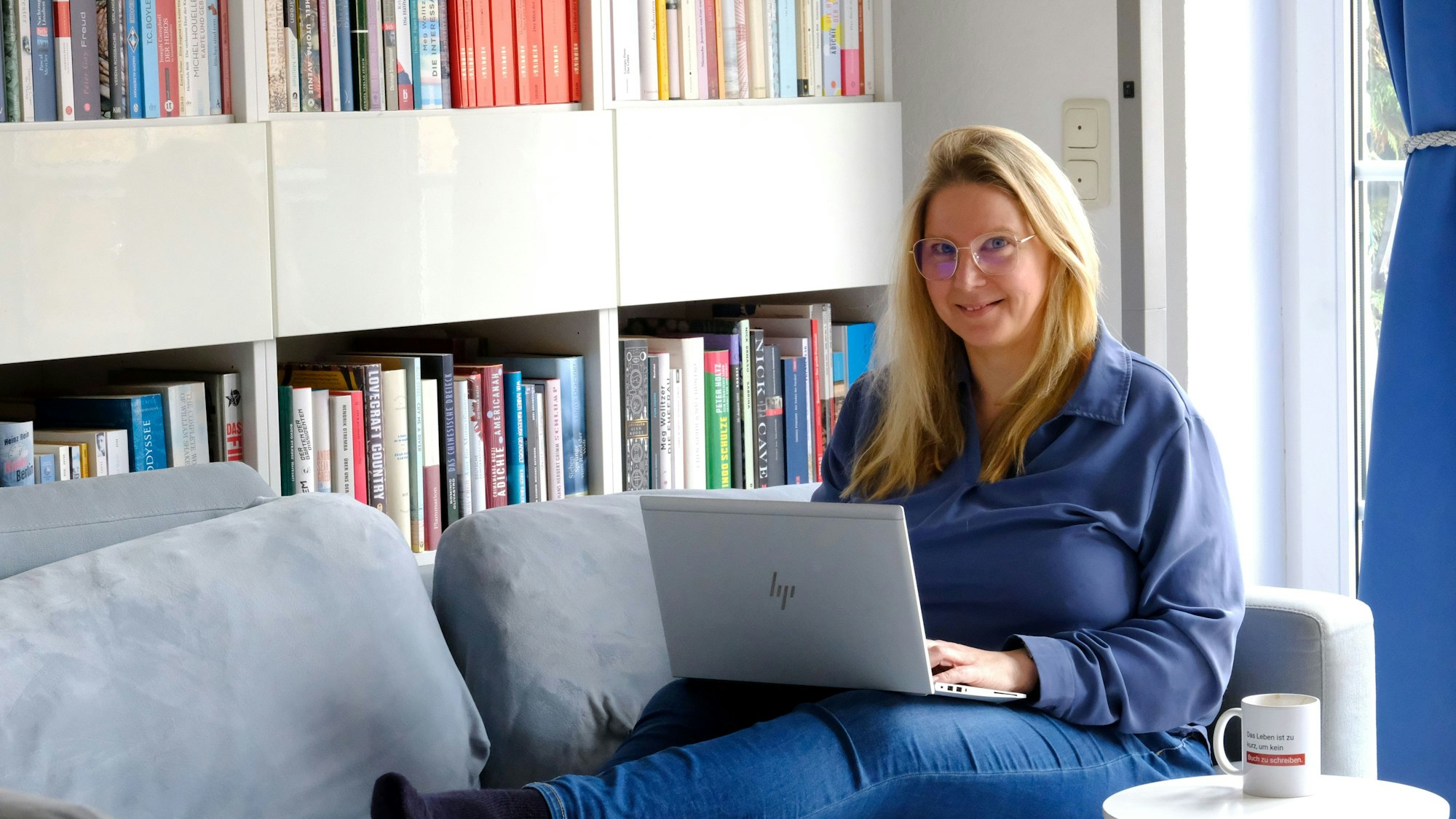 Yvonne Kraus aus Eicks, die als Buchcoach arbeitet, sitzt mit ihrem Laptop auf dem Sofa vor einem Bücherregal.