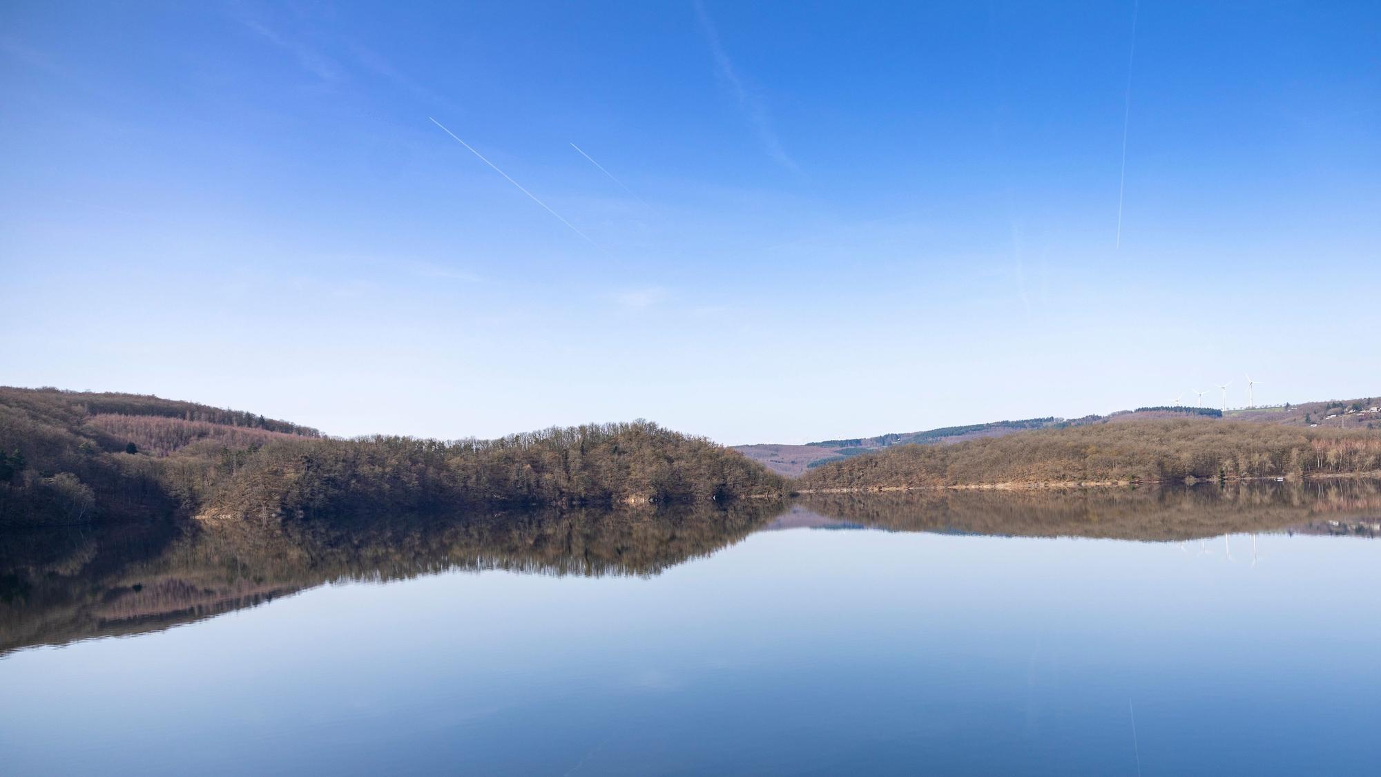 Blick auf den stillen Rursee, in dem sich der blaue Himmel spiegelt.