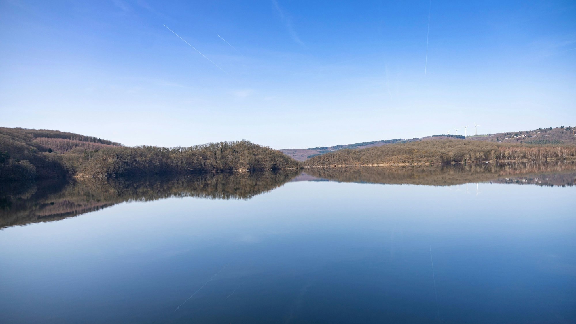 Blick auf den stillen Rursee, in dem sich der blaue Himmel spiegelt.