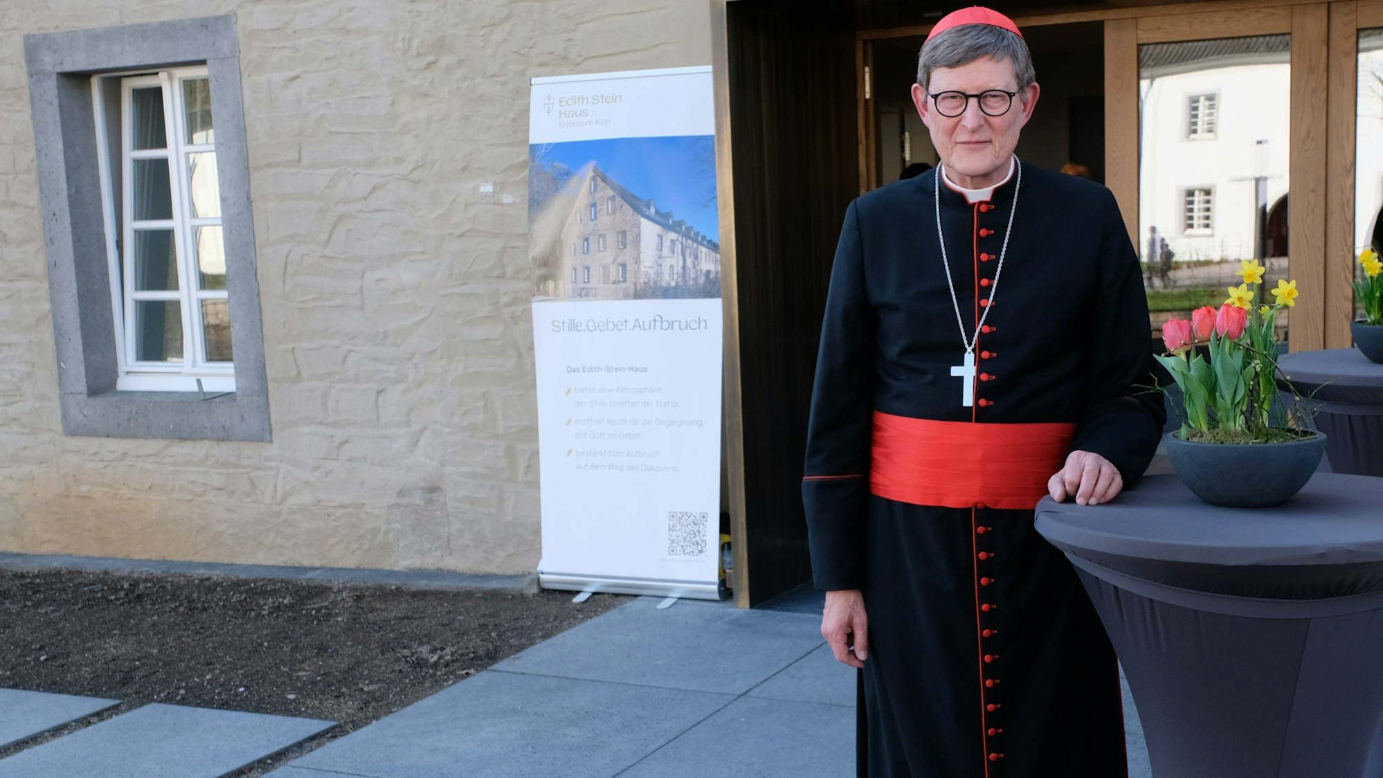 Ein Kardinal in schwarz-purpur Talar steht vor einem Haus.