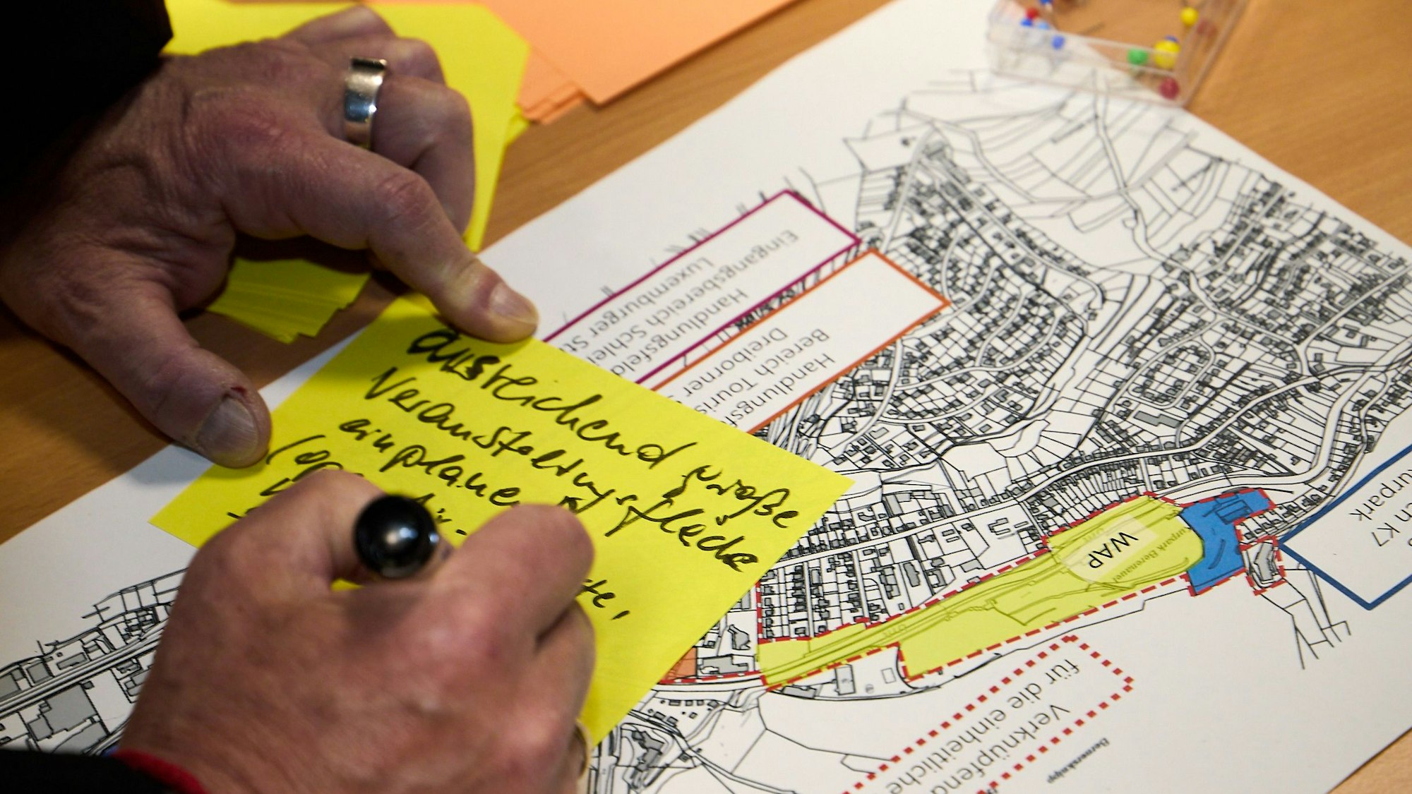 Auf einem Plan liegt ein gelber Zettel, auf den jemand gerade etwas schreibt.