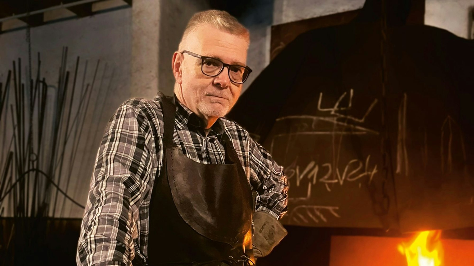 Ralf Lano steht vor einem Schmiedeofen und trägt Schutzkleidung. Er stützt sich auf einen Hammer im Bildvordergrund, der auf einer Fläche auf Hüfthöhe aufliegt.