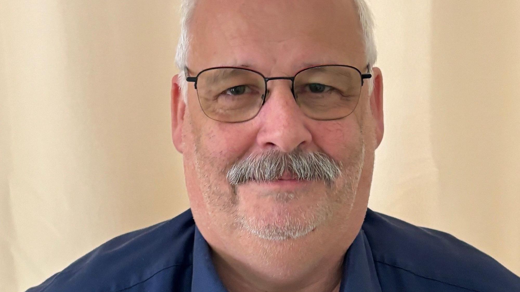 Auf dem Bild ist das Portrait eines Mannes mit Brille und in einem blauen Hemd zu sehen.