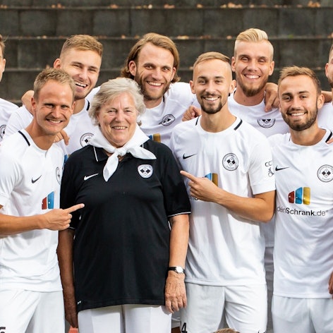 Auf dem Bild ist eine ältere Dame im Trikot zu sehen, die von Fußballspielern umringt ist.
