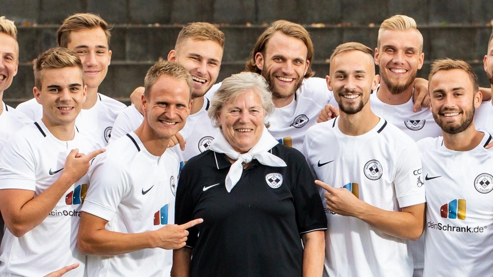 Auf dem Bild ist eine ältere Dame im Trikot zu sehen, die von Fußballspielern umringt ist.