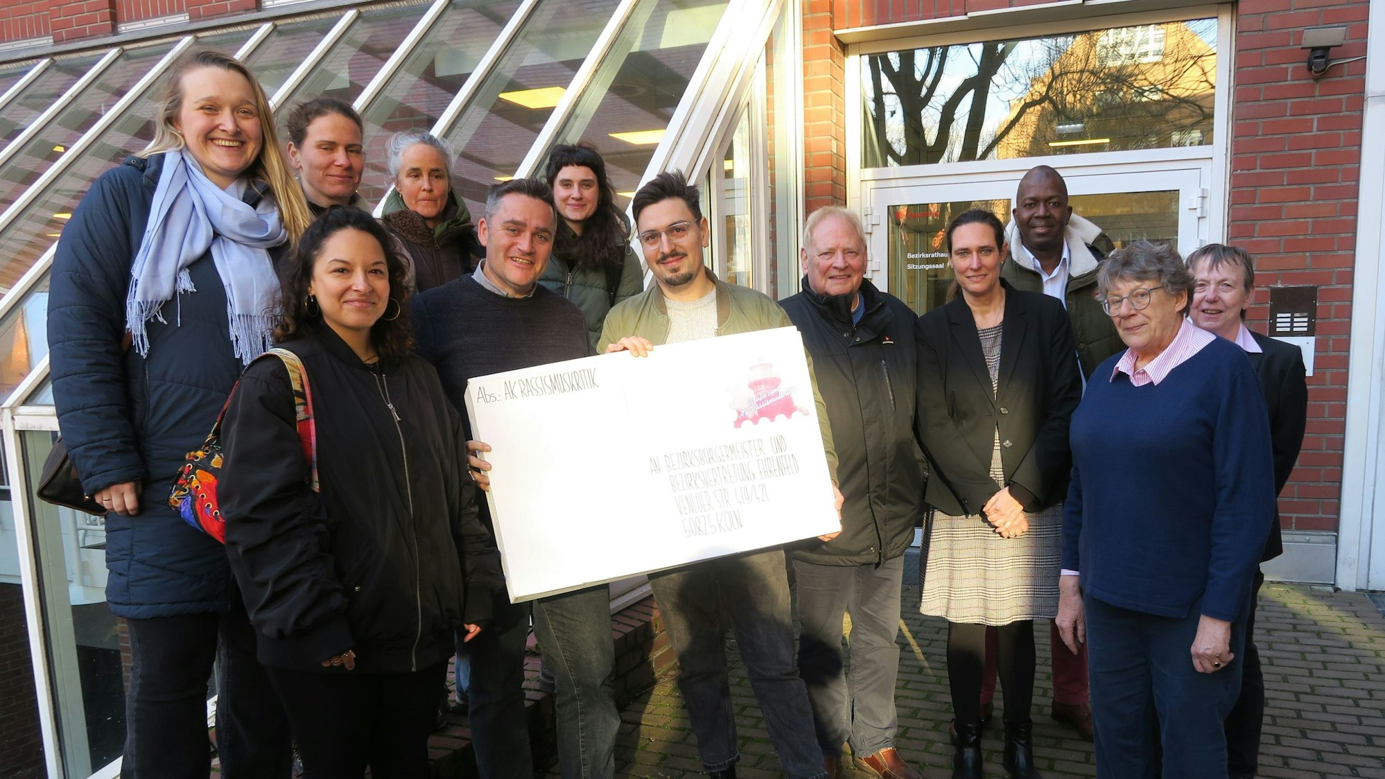 Zwölf Personen stehen nebeneinander. Zwei Männer in der Mitte halten einen übergroßen Briefumschlag in den Händen.