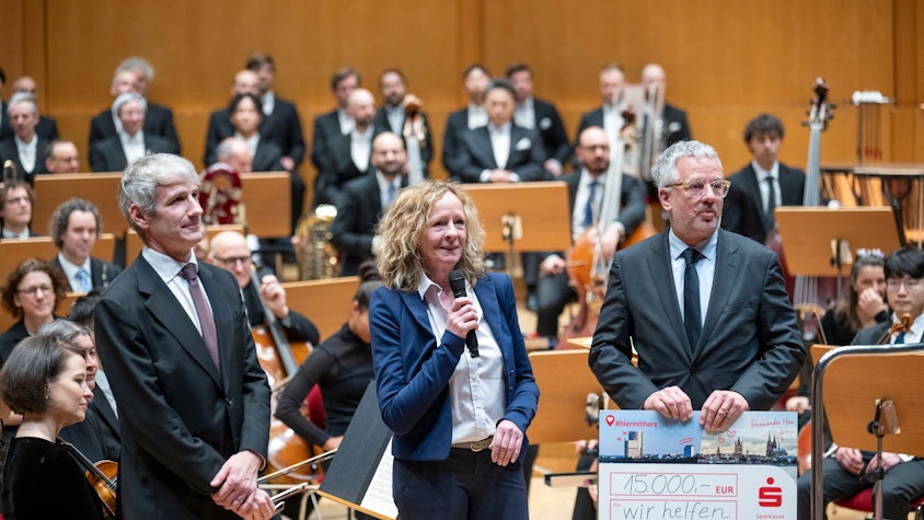 Isabella NevenDuMont und Christian Hümmeler stehen auf der Bühne der Kölner Philharmonie und bedanken sich im Namen von „wir helfen“ bei Gürzenich-Chef Stefan Englert und seinem Orchester für die Spende in Höhe von 15.000 Euro