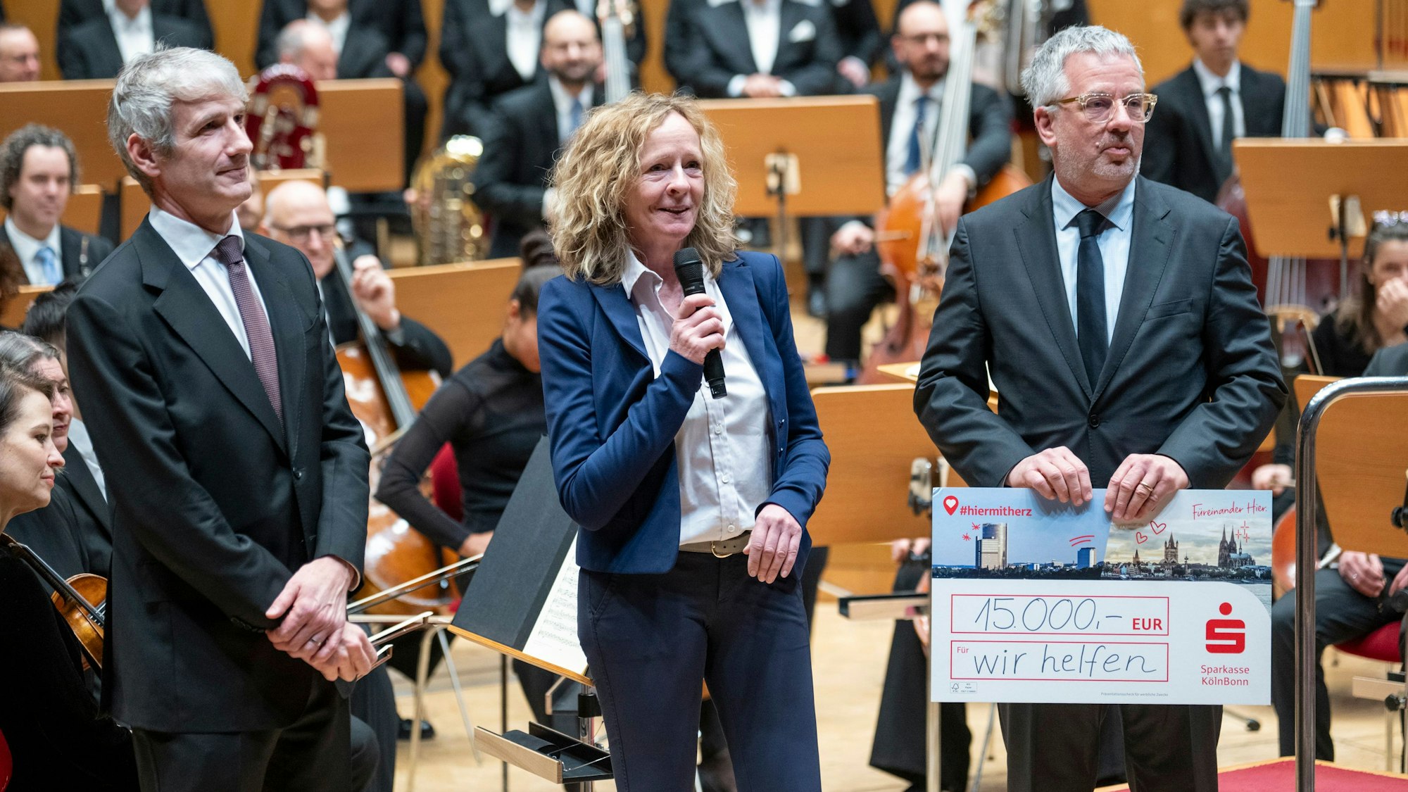 Isabella NevenDuMont und Christian Hümmeler stehen auf der Bühne der Kölner Philharmonie und bedanken sich im Namen von „wir helfen“ bei Gürzenich-Chef Stefan Englert und seinem Orchester für die Spende in Höhe von 15.000 Euro