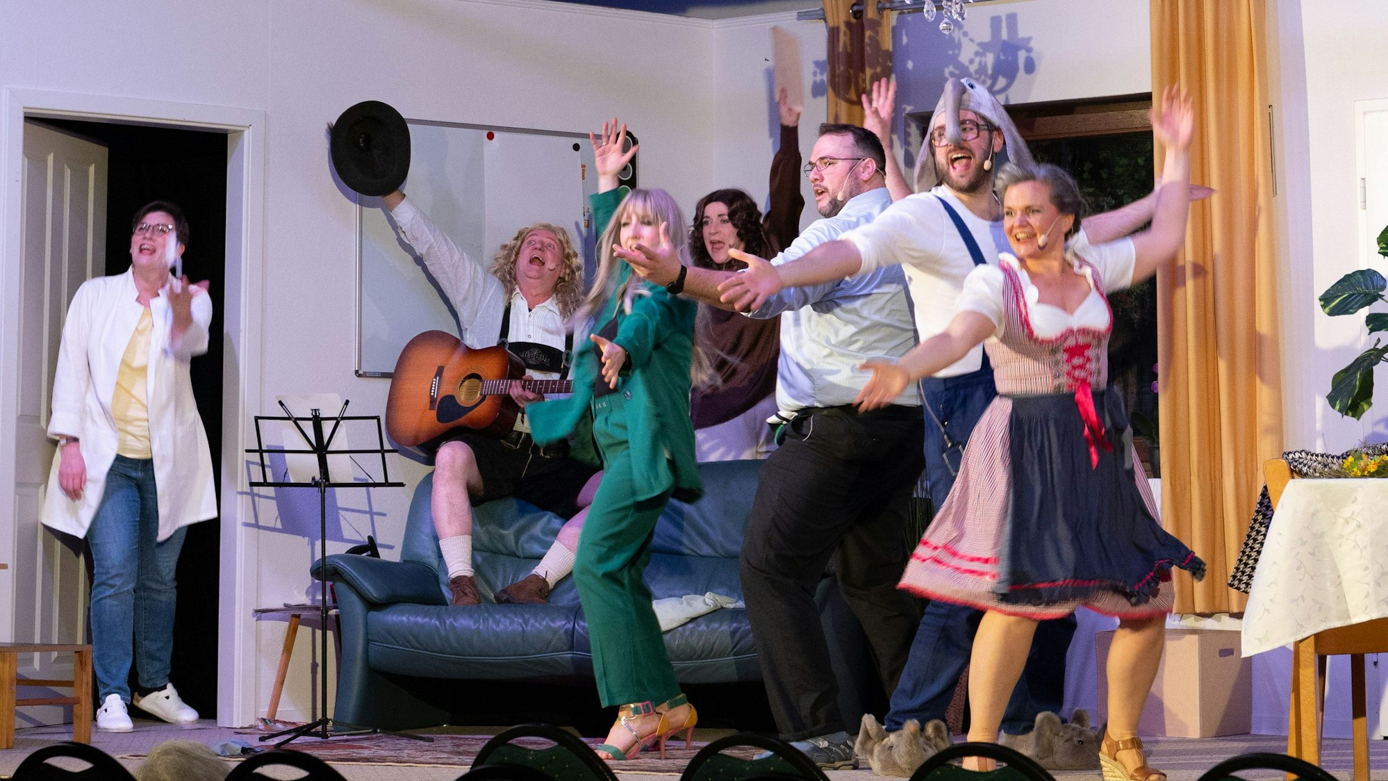 Mehrere Menschen stehen tanzend und singend auf einer Bühne. Das Bühnenbild zeigt ein Wohnzimmer.