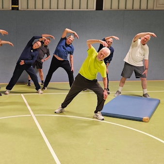 Eine Gruppe von älteren Männern macht Dehnübungen in einer Sporthalle.