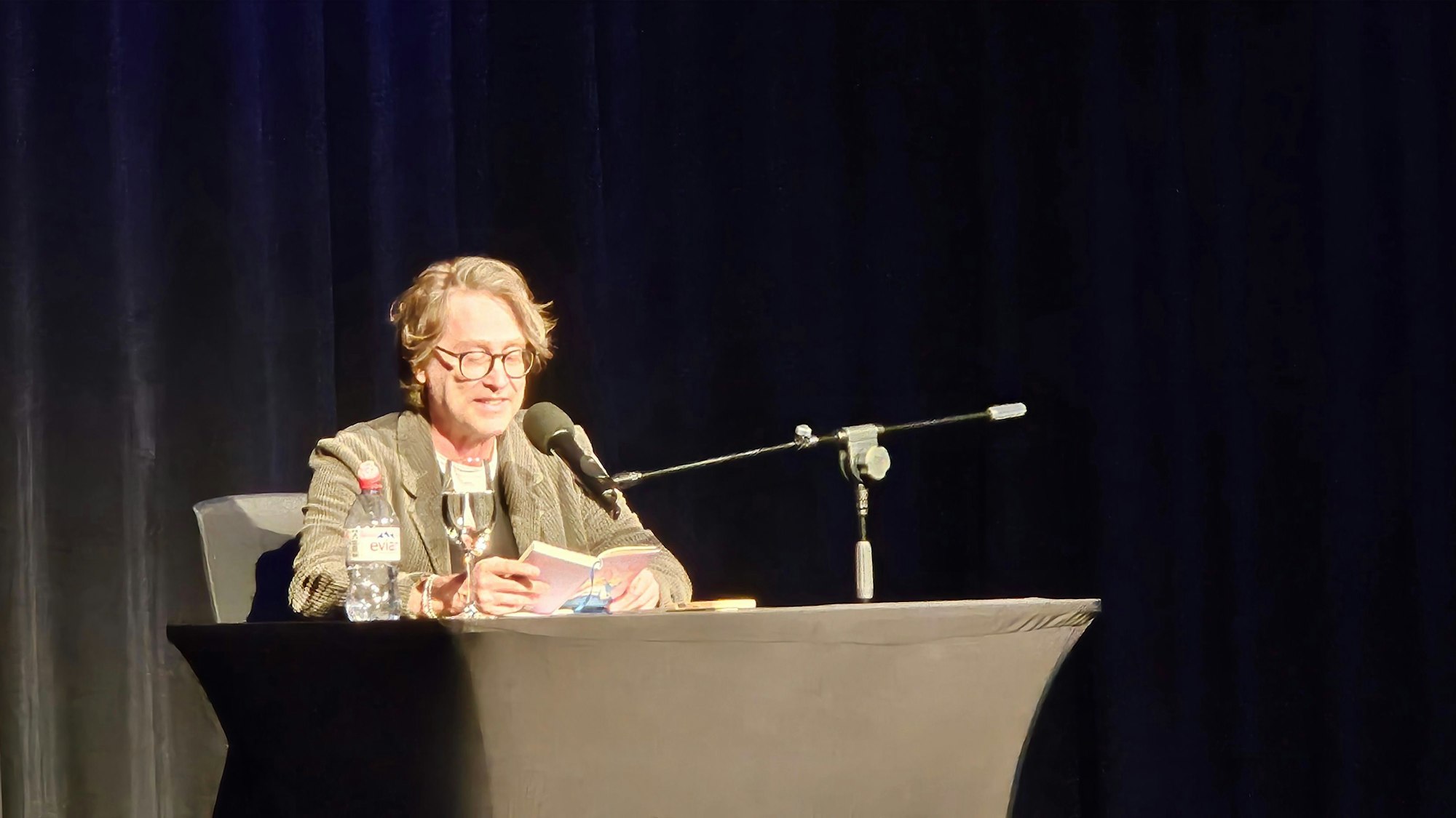 Das Bild zeigt den Autor sitzend auf der Bühne, er hat sein Buch aufgeschlagen und liest daraus, vor ihm ein Mikrofon, ein Glas und eine Flasche Wasser.