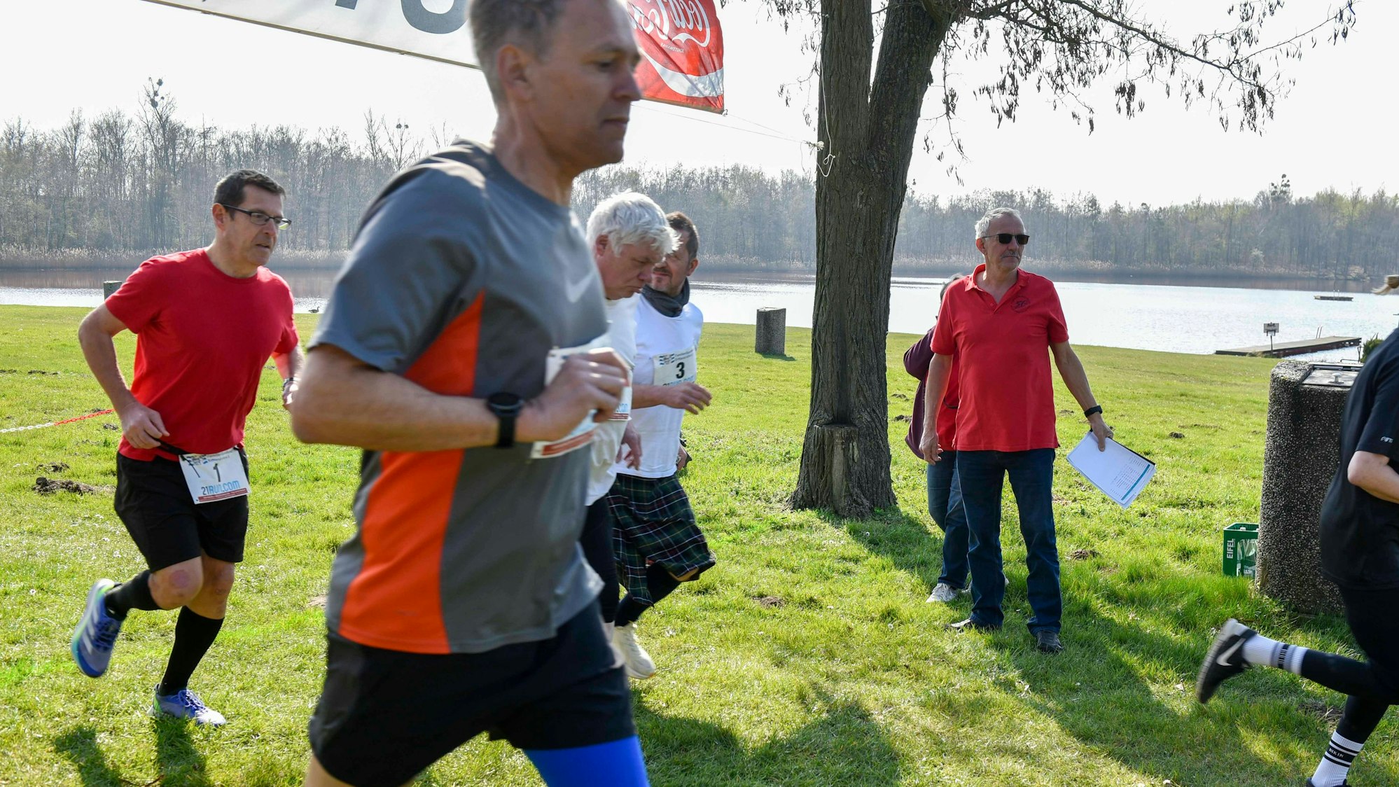 Zu sehen sind mehrere Läufer auf Rasen an einem See.