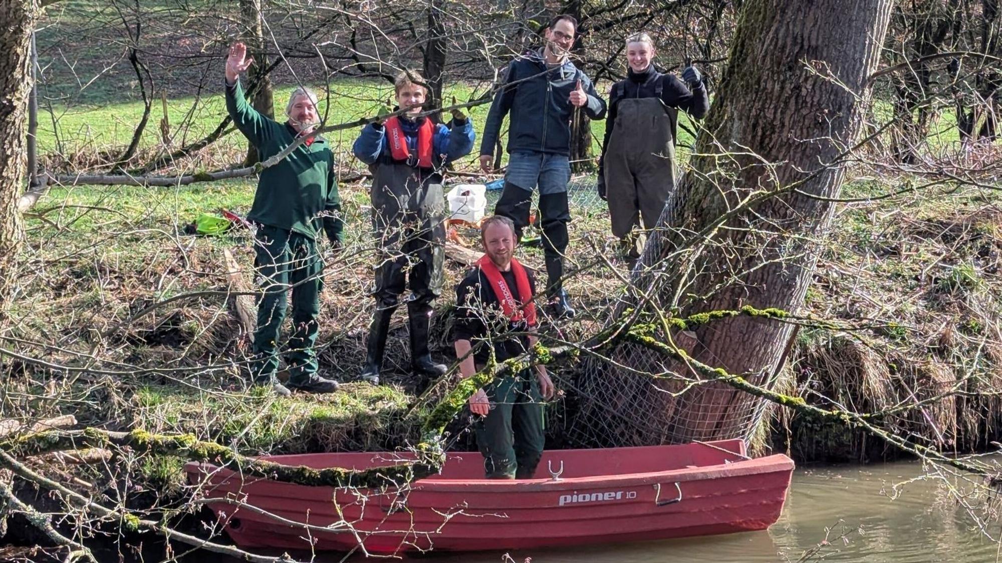 Ein rotes Boot auf dem Wasser, darin ein Mann. Am Ufer vier weitere Personen neben einem Baum, der über der Wasseroberfläche circa einen Meter hoch mit Metall umwickelt ist.