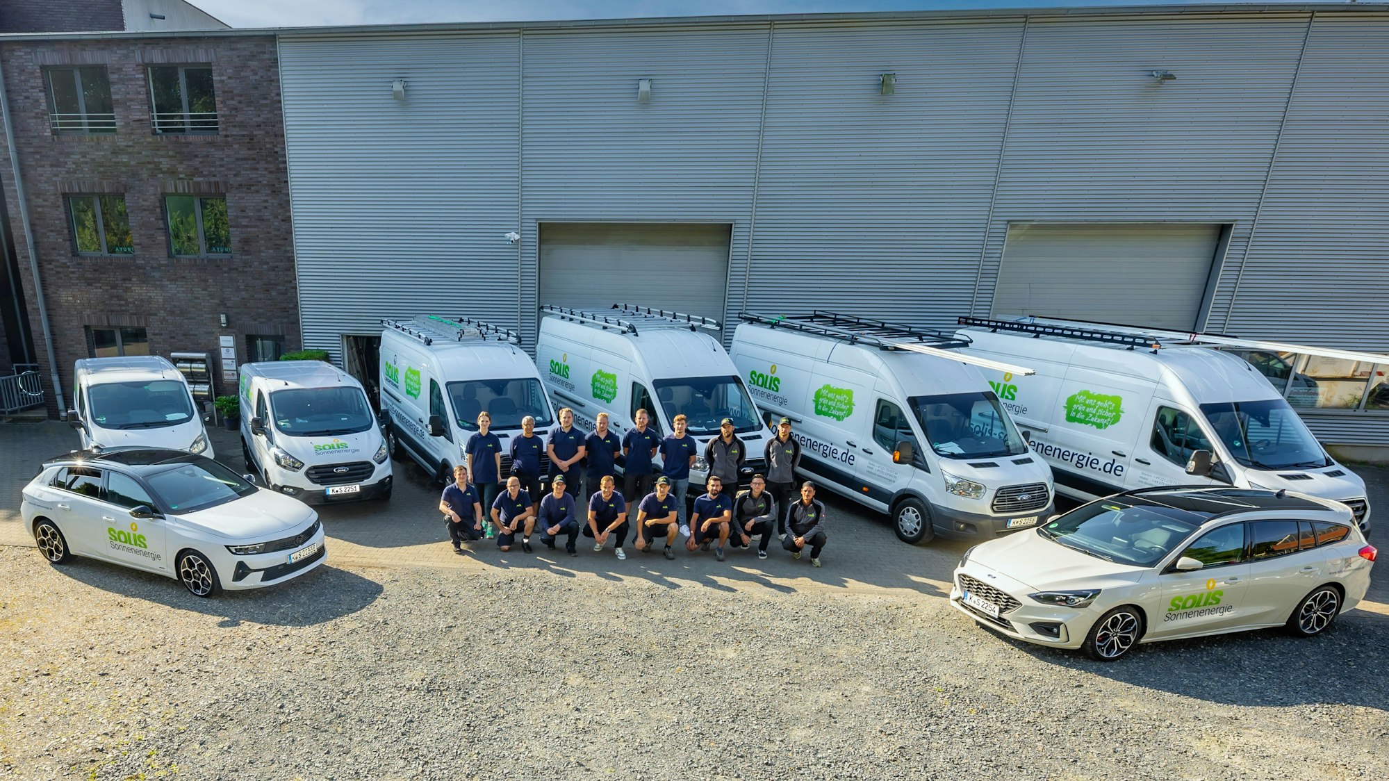 Auf dem Hof der Firma stehen mehrere gelabelte Autos des Unternehmens Solis Sonnenenergie. Die Mitarbeiter stehen in der Mitte zum Gruppenfoto in zwei Reihen aufgereiht.