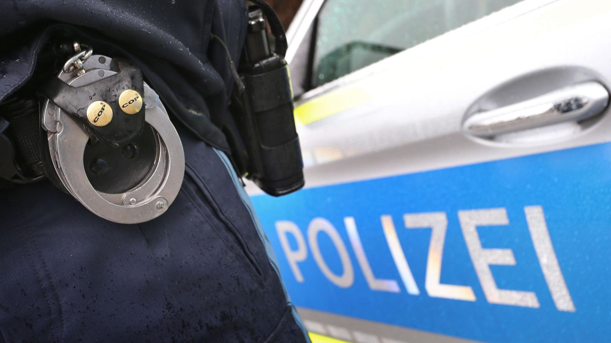 Ein Polizist mit Handschellen und einer Pistole am Gürtel steht vor einem Streifenwagen.